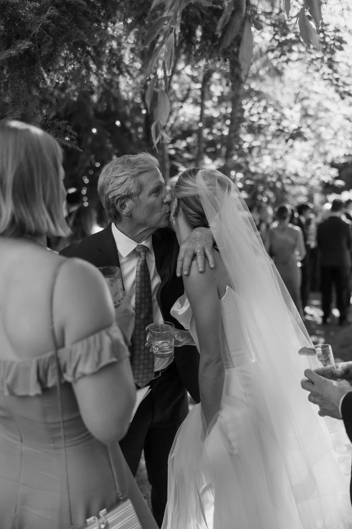 A father embracing and kissing the bride, surrounded by family on her wedding day.